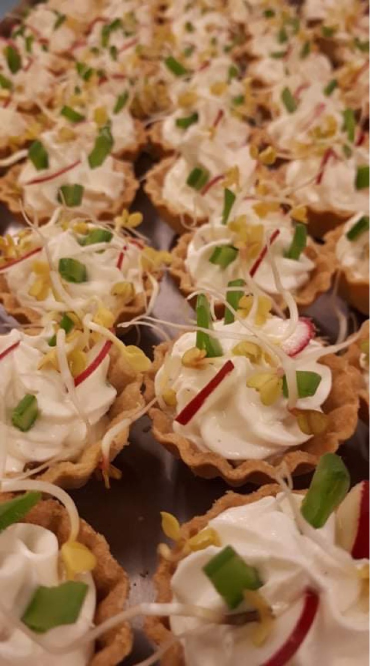 Kruche babeczki wypełnione kremowym serem, udekorowane rzodkiewką, szczypiorkiem i kiełkami, ułożone na ciemnej tacy, widok z góry pod kątem.