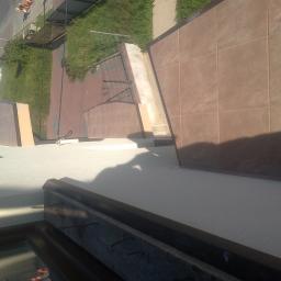 Zakład remontowo-budowlany - Widok z góry na wyłożony brązowymi płytkami taras, z widocznym fragmentem okna i elementami konstrukcyjnymi, oraz zarysem cienia osoby fotografującej.