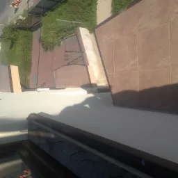 Widok z góry na wyłożony brązowymi płytkami taras, z widocznym fragmentem okna i elementami konstrukcyjnymi, oraz zarysem cienia osoby fotografującej.