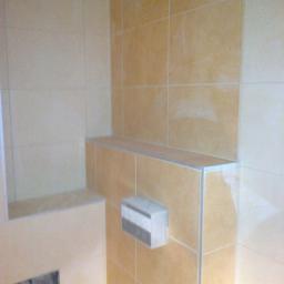 Zakład remontowo-budowlany - Beżowe płytki ceramiczne na ścianie łazienki, widoczny fragment zabudowy z półką i przyciskiem spłuczki, listwy wykończeniowe na krawędziach.