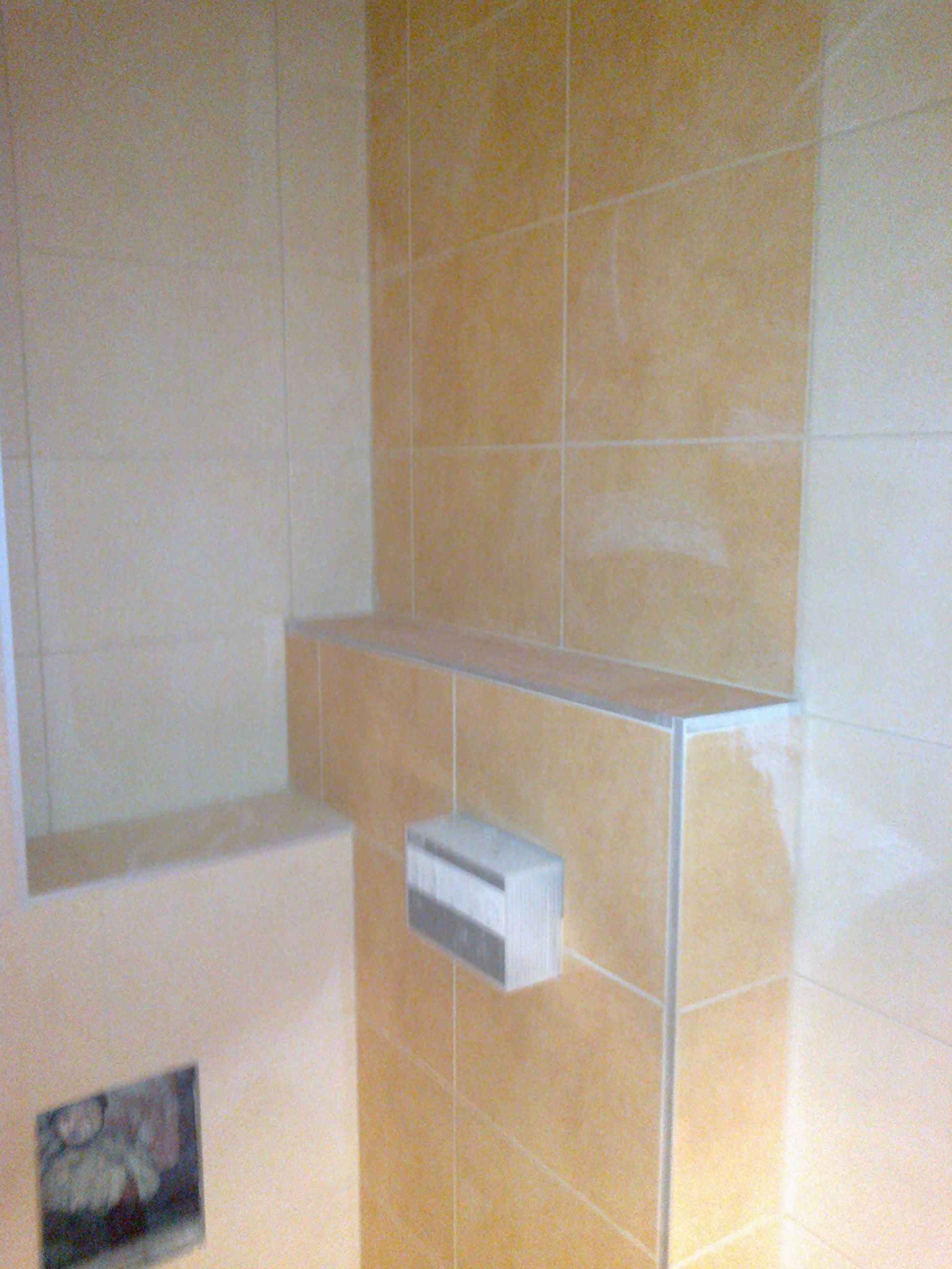 Beżowe płytki ceramiczne na ścianie łazienki, widoczny fragment zabudowy z półką i przyciskiem spłuczki, listwy wykończeniowe na krawędziach.