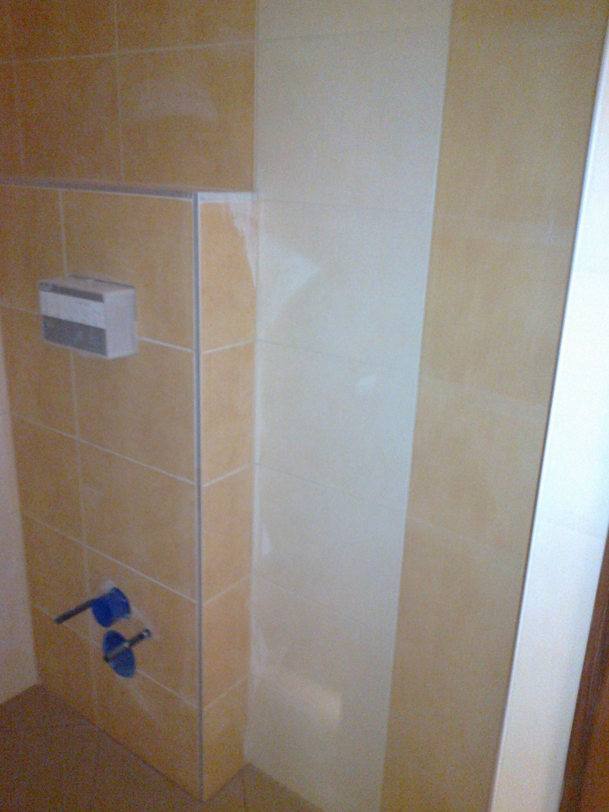 Fragment ściany w łazience z częściowo położonymi, beżowymi płytkami, widoczne wystające elementy instalacji wodnej zabezpieczone niebieską folią oraz podajnik na mydło w płynie.