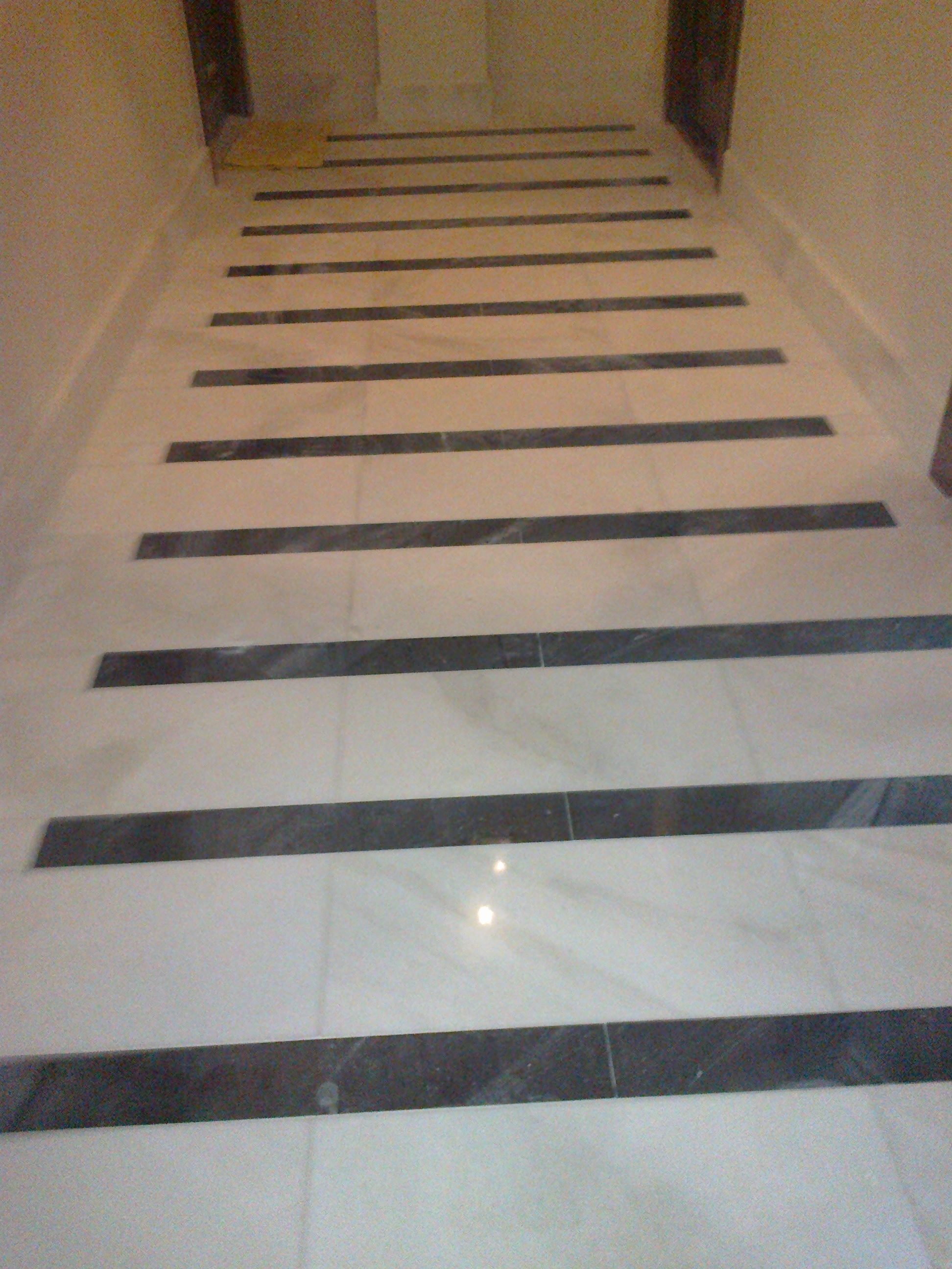 Biała marmurowa podłoga z wstawkami z ciemnego kamienia ułożonymi w równych odstępach, tworzącymi wzór pasów w korytarzu.