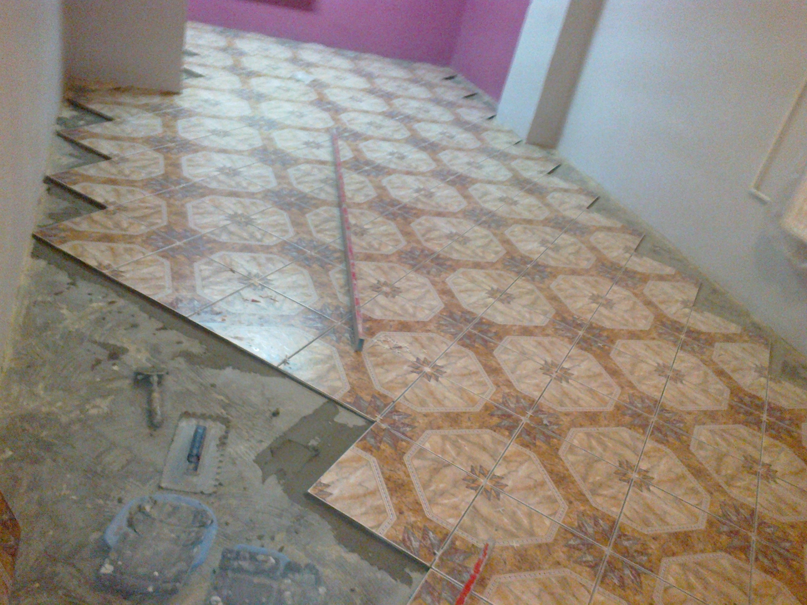 Układanie brązowo-beżowych płytek podłogowych z wzorem kwiatowym, widoczne narzędzia glazurnicze i fragment przygotowanej podłogi w trakcie prac remontowych.