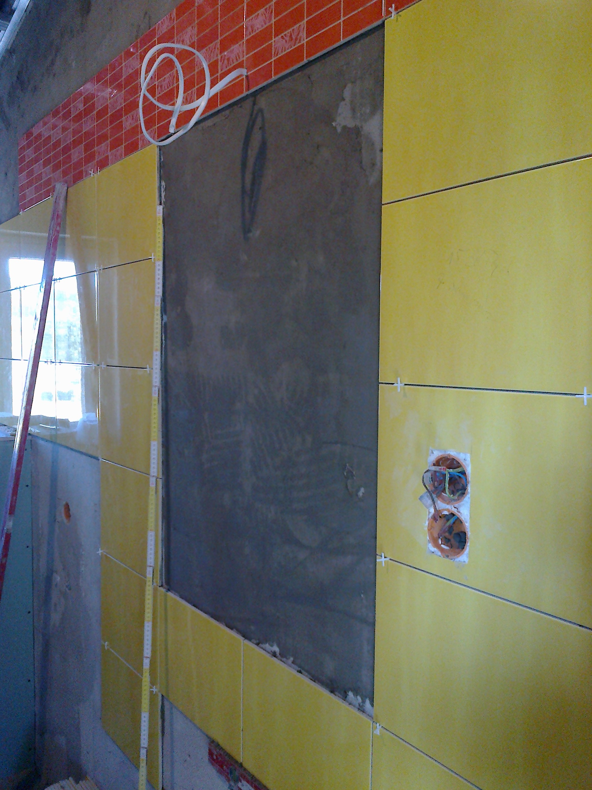 Żółte i czerwone płytki ułożone na ścianie, widoczne krzyżyki dystansowe, poziomica i otwory pod gniazdka elektryczne, etap prac wykończeniowych.