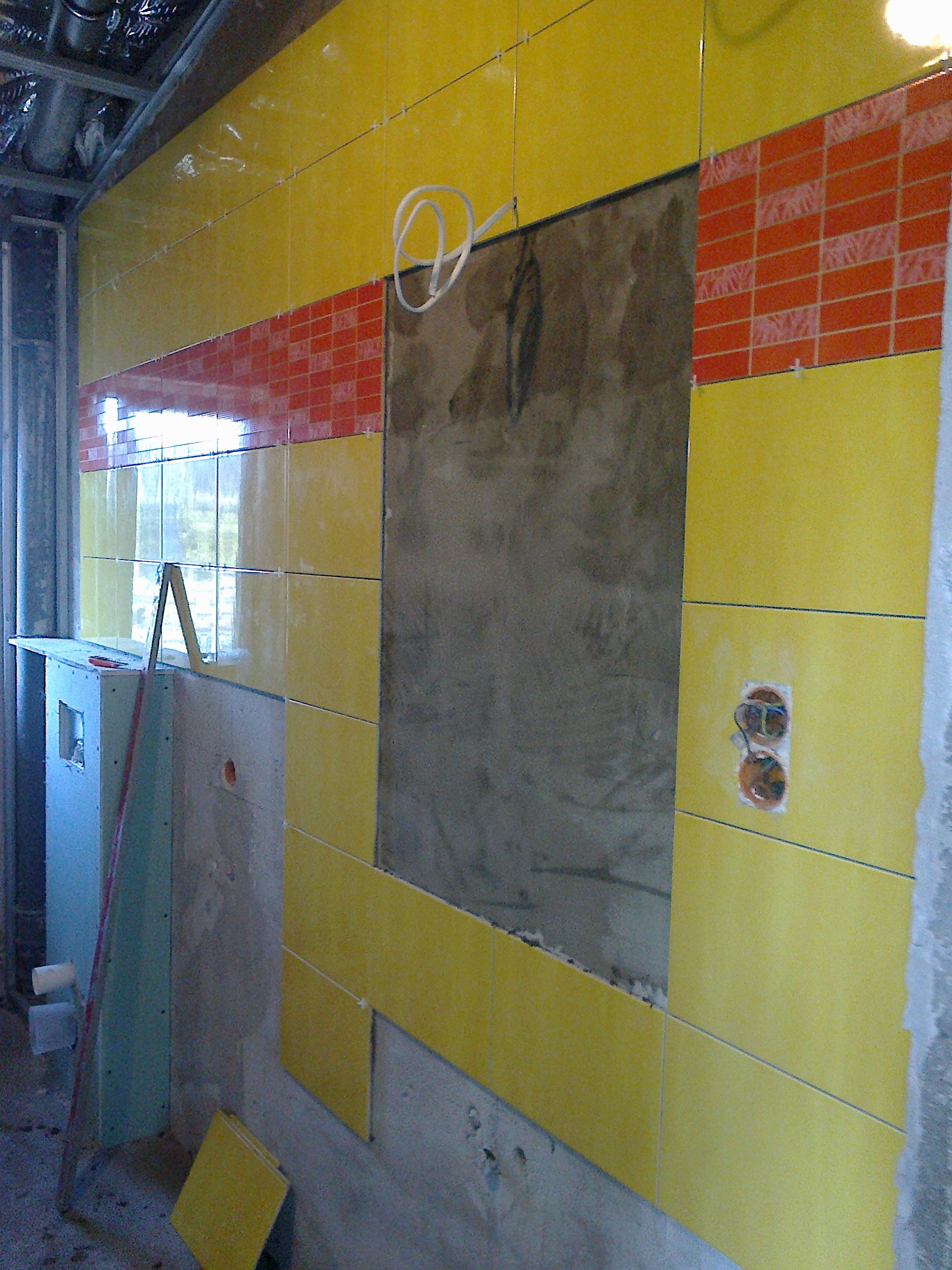 Żółto-pomarańczowa okładzina ścienna w trakcie montażu, widoczne krzyżyki dystansowe, fragment ściany bez płytek i wystające kable elektryczne.