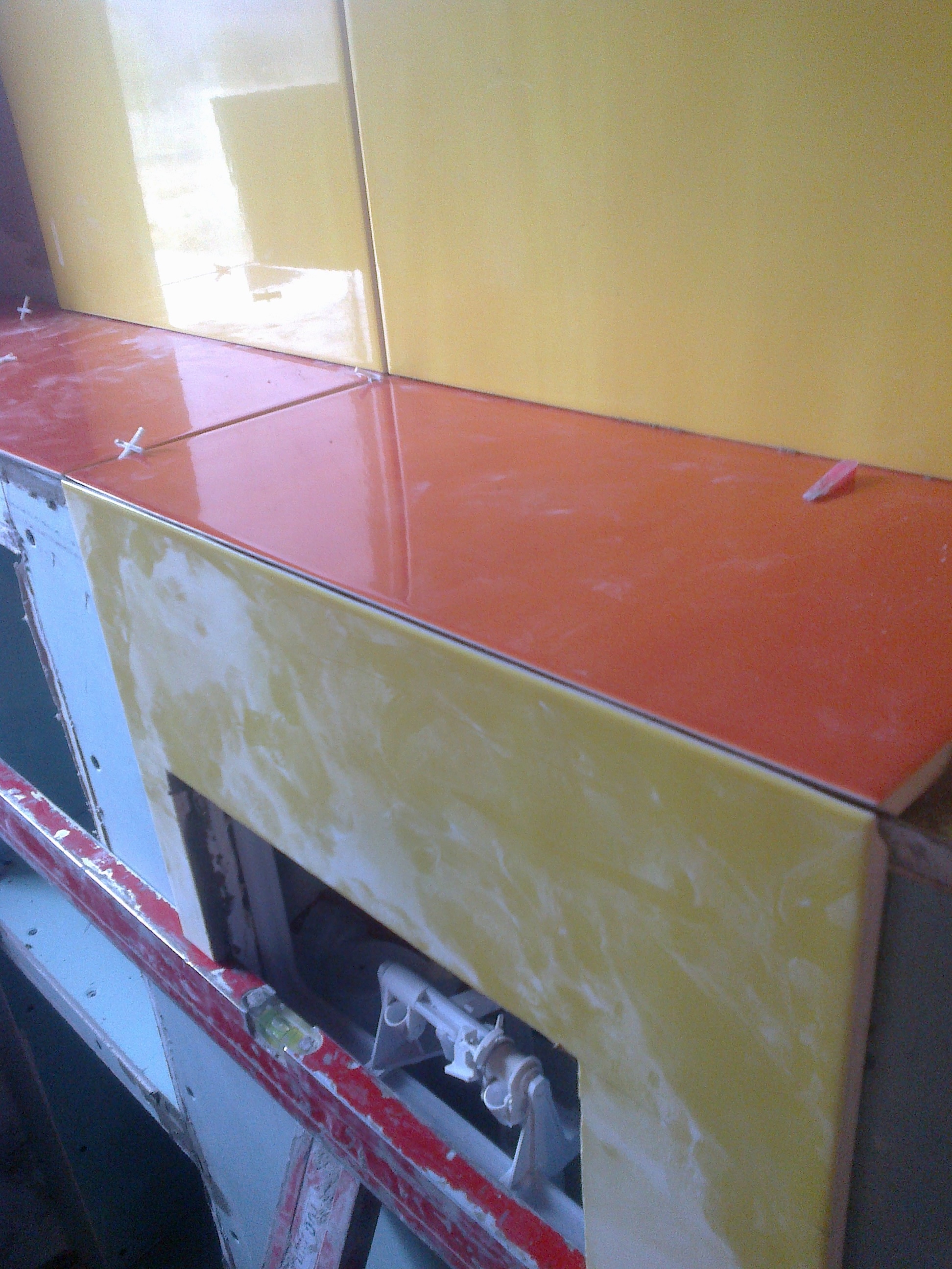 Montaż pomarańczowych i żółtych płytek ceramicznych w łazience, widoczne krzyżyki dystansowe, poziomica i elementy instalacji podtynkowej.