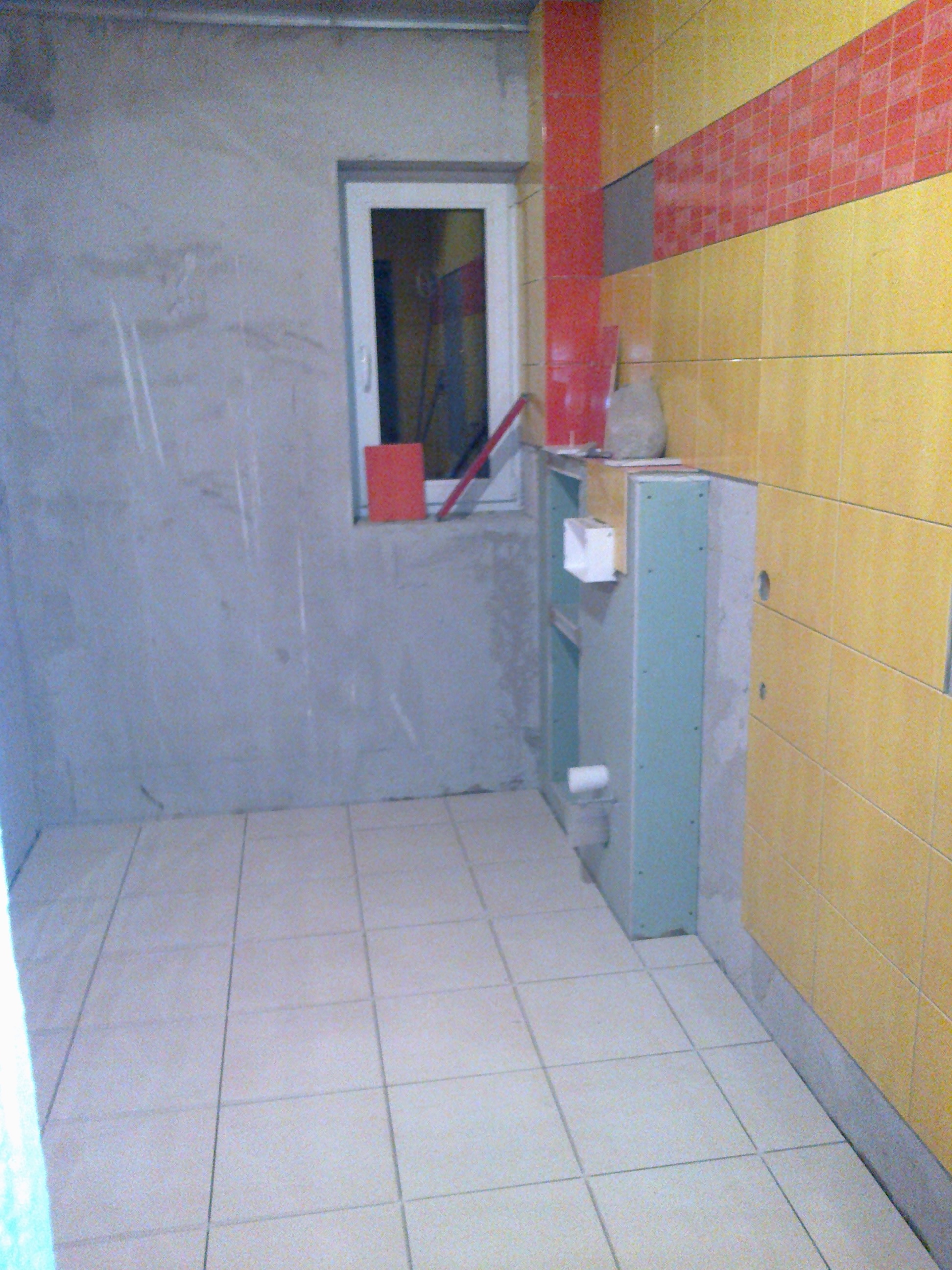 Wnętrze łazienki w trakcie remontu: ułożone jasne płytki podłogowe, częściowo wykończone ściany z żółto-czerwonymi kafelkami, obudowa stelaża podtynkowego WC z płyt karton-gips, surowa ściana...
