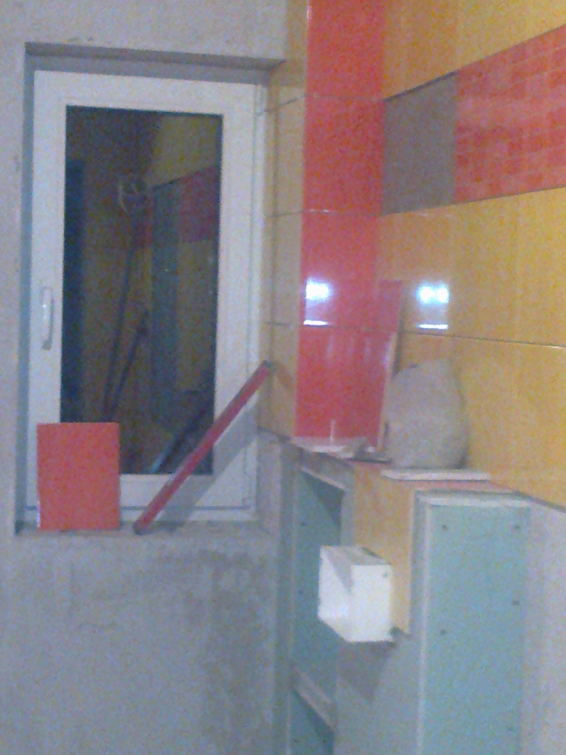 Fragment remontowanej kuchni z widocznymi płytkami w odcieniach czerwieni i żółci, surowymi ścianami, oknem z parapetem i narzędziami, ukazujący etap przygotowania do dalszych prac wykończeniowych.