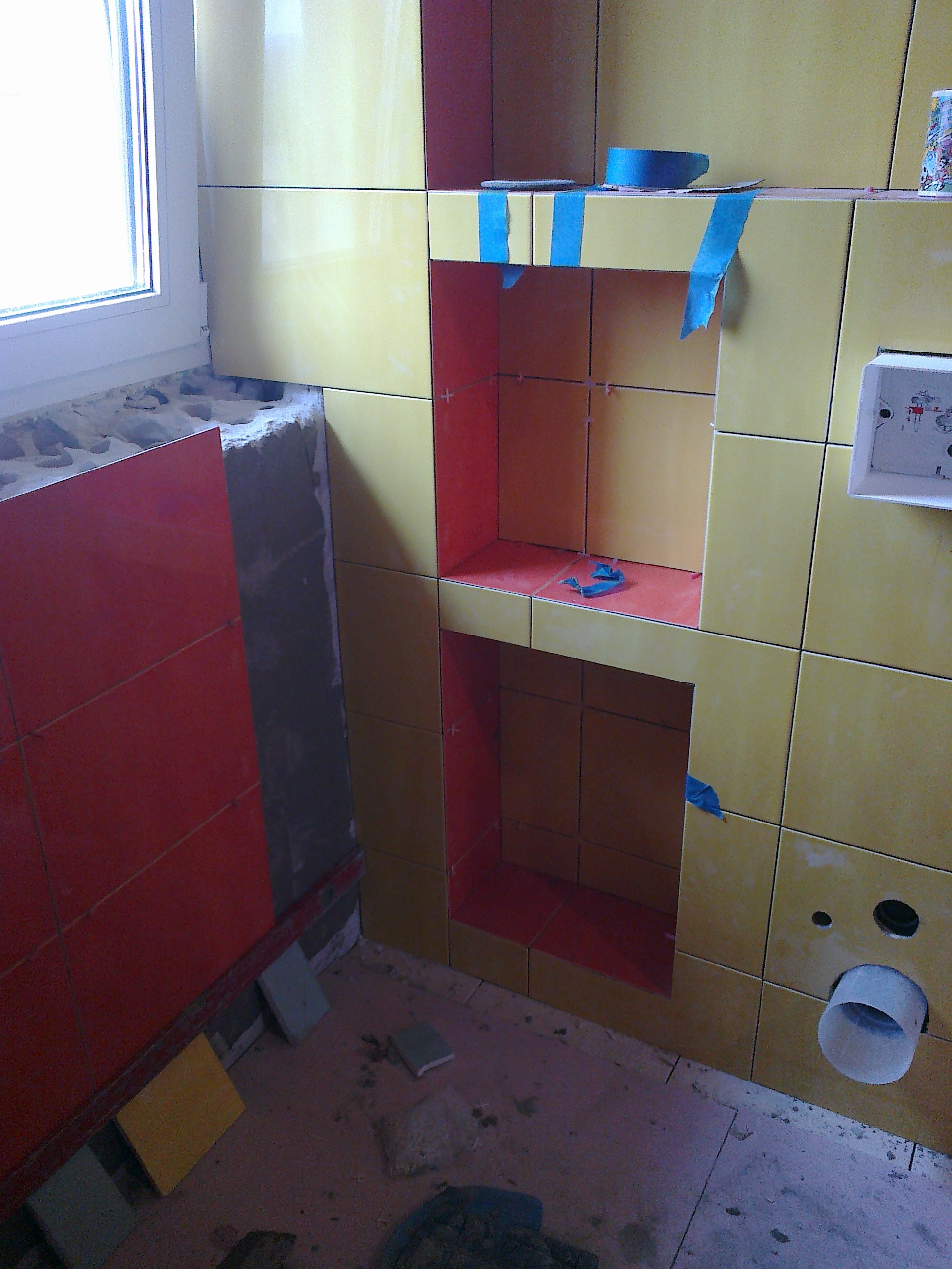Żółto-czerwona mozaika ścienna z wnękami w trakcie montażu, zabezpieczona niebieską taśmą malarską, widoczny otwór kanalizacyjny i fragment okna.