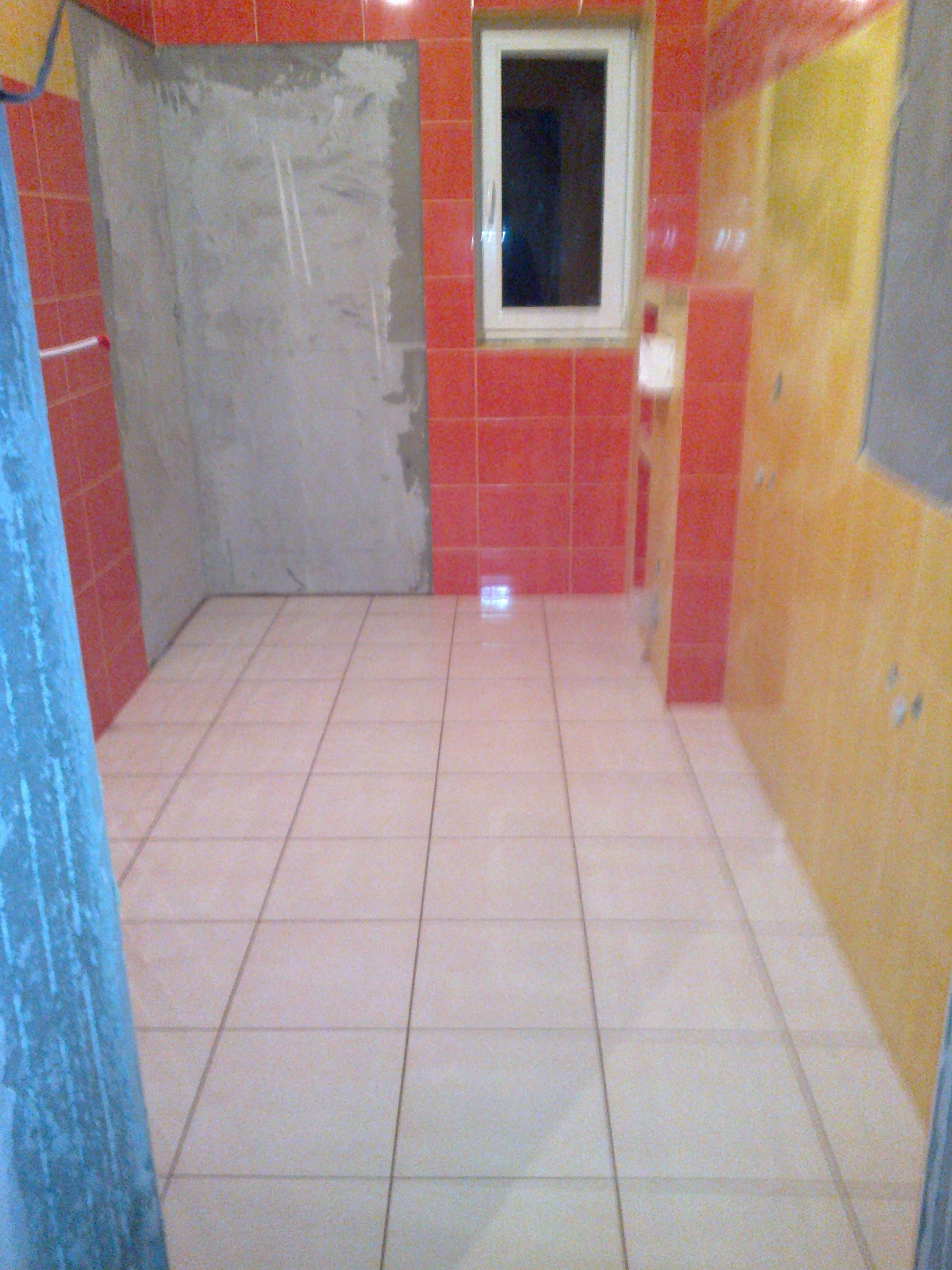 Wnętrze łazienki w trakcie remontu, widoczne położone płytki podłogowe, częściowo wykończone ściany w czerwonej i żółtej glazurze, oraz surowy, nieotynkowany fragment ściany obok okna.