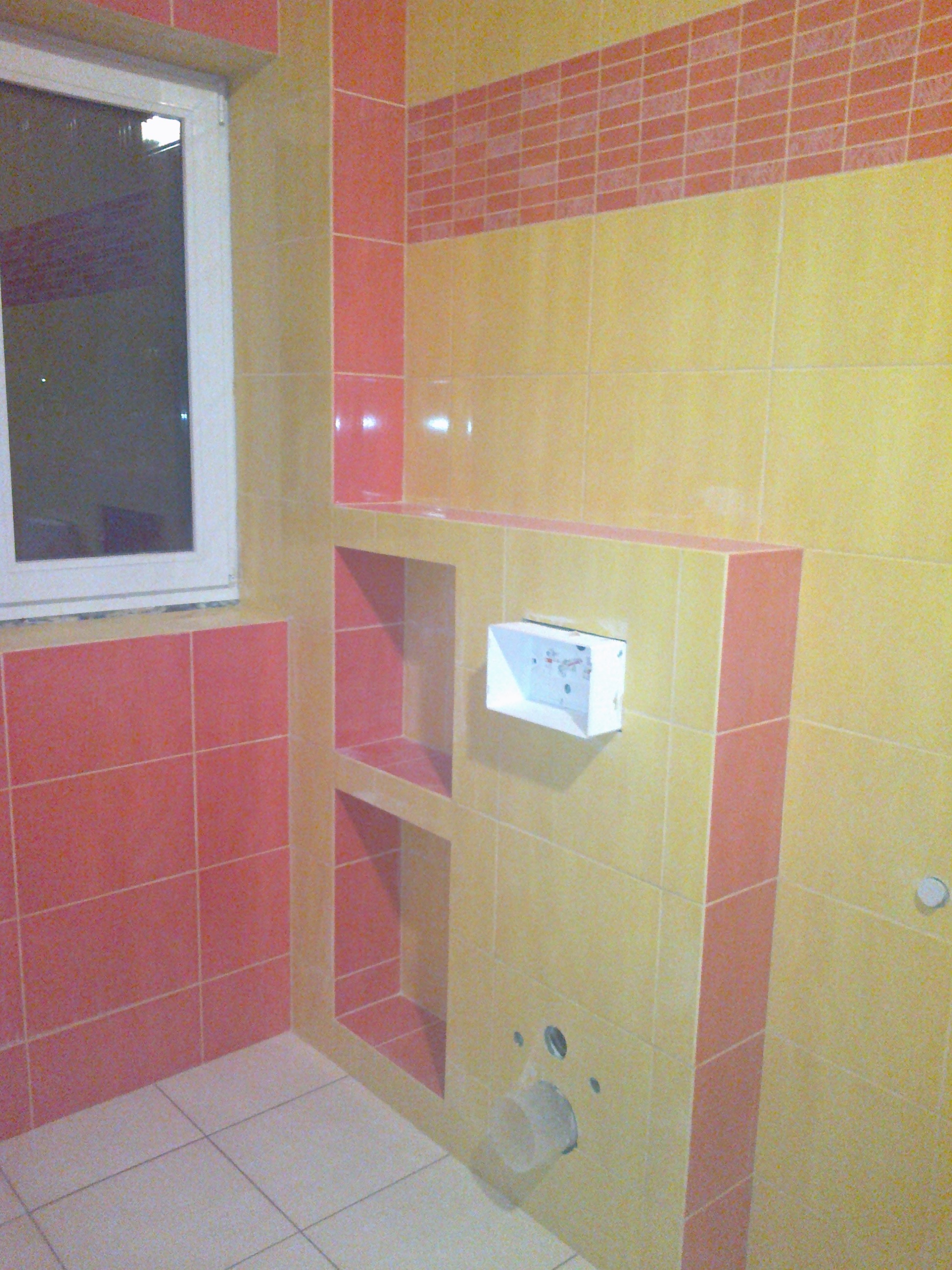 Wykończona łazienka z żółto-różowymi płytkami, wnękami w ścianie i przygotowanym miejscem na armaturę.