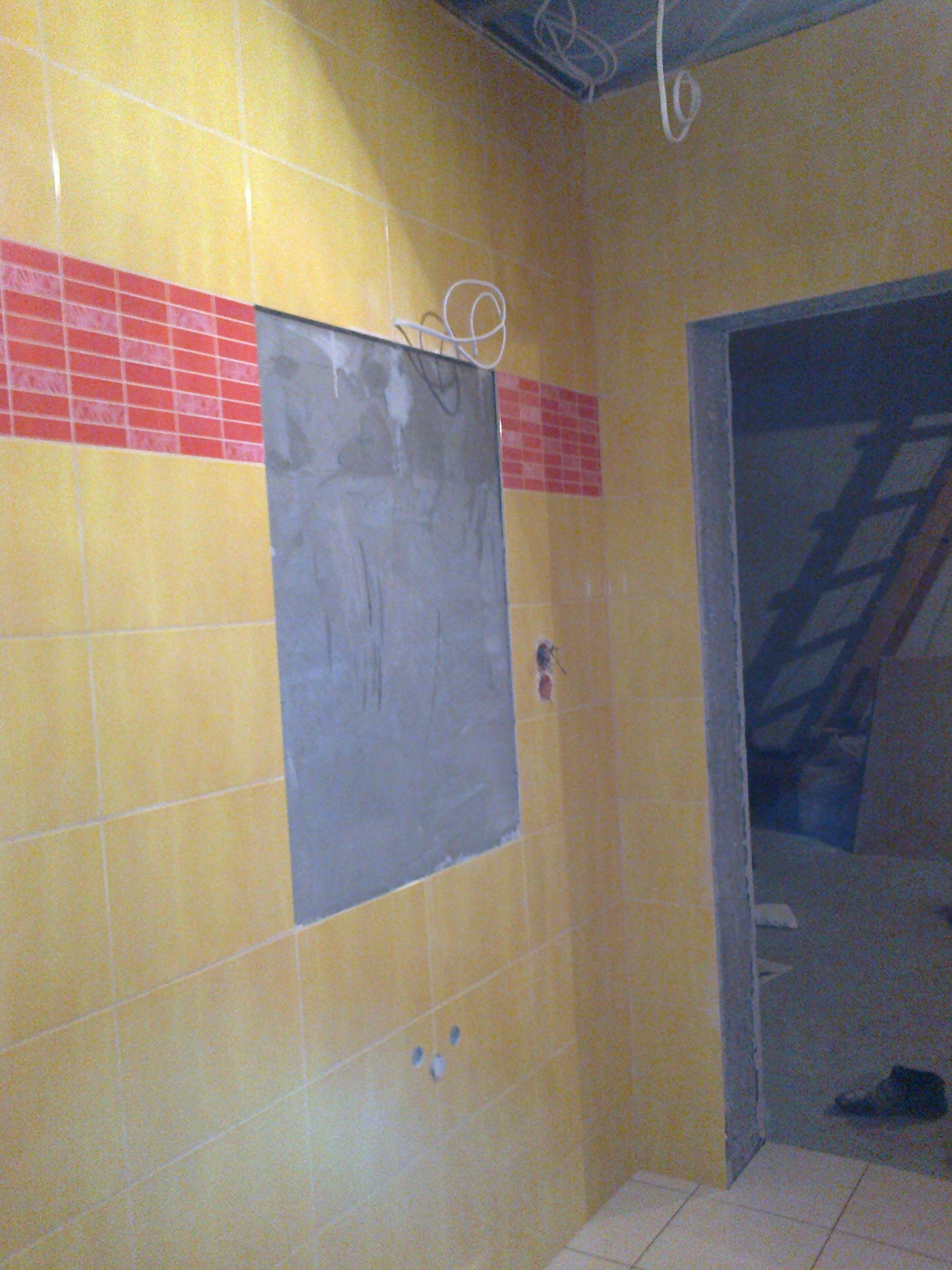 Żółte płytki ścienne z czerwonym pasem dekoracyjnym w trakcie układania, widoczne niedokończone miejsce na lustro i wystające przewody elektryczne w łazience w trakcie remontu.