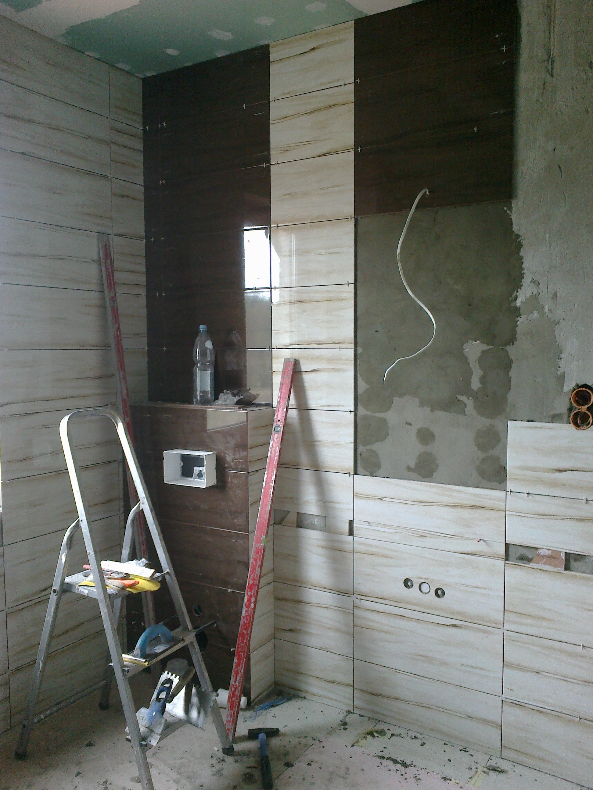Proces układania płytek imitujących drewno w łazience, widoczne narzędzia, poziomica oparta o ścianę, drabina z narzędziami, częściowo wykończona ściana z widocznymi otworami na instalacje.