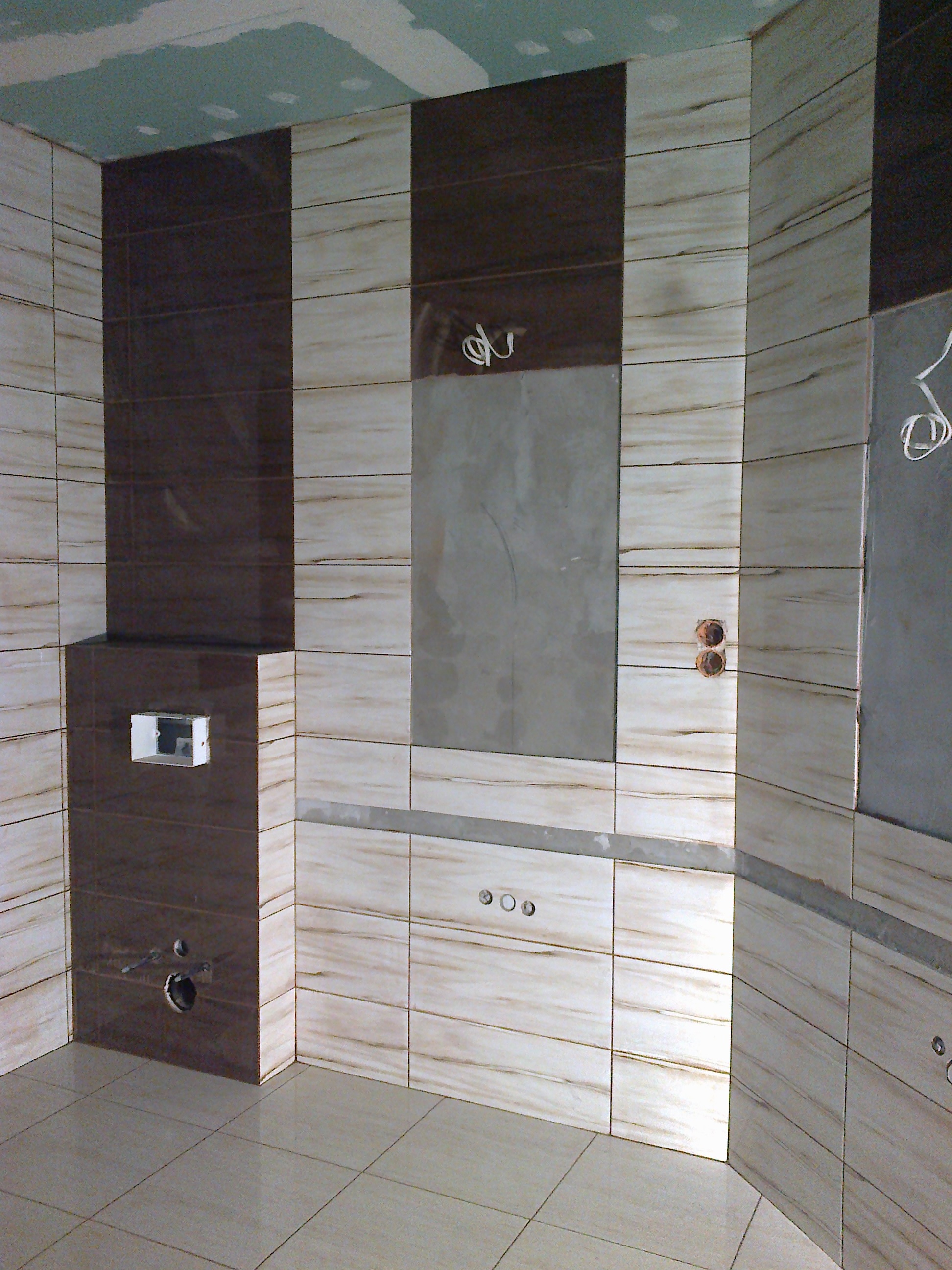 Wnętrze łazienki w trakcie remontu, widoczne płytki imitujące drewno w odcieniach beżu i brązu, surowy sufit i instalacje elektryczne.