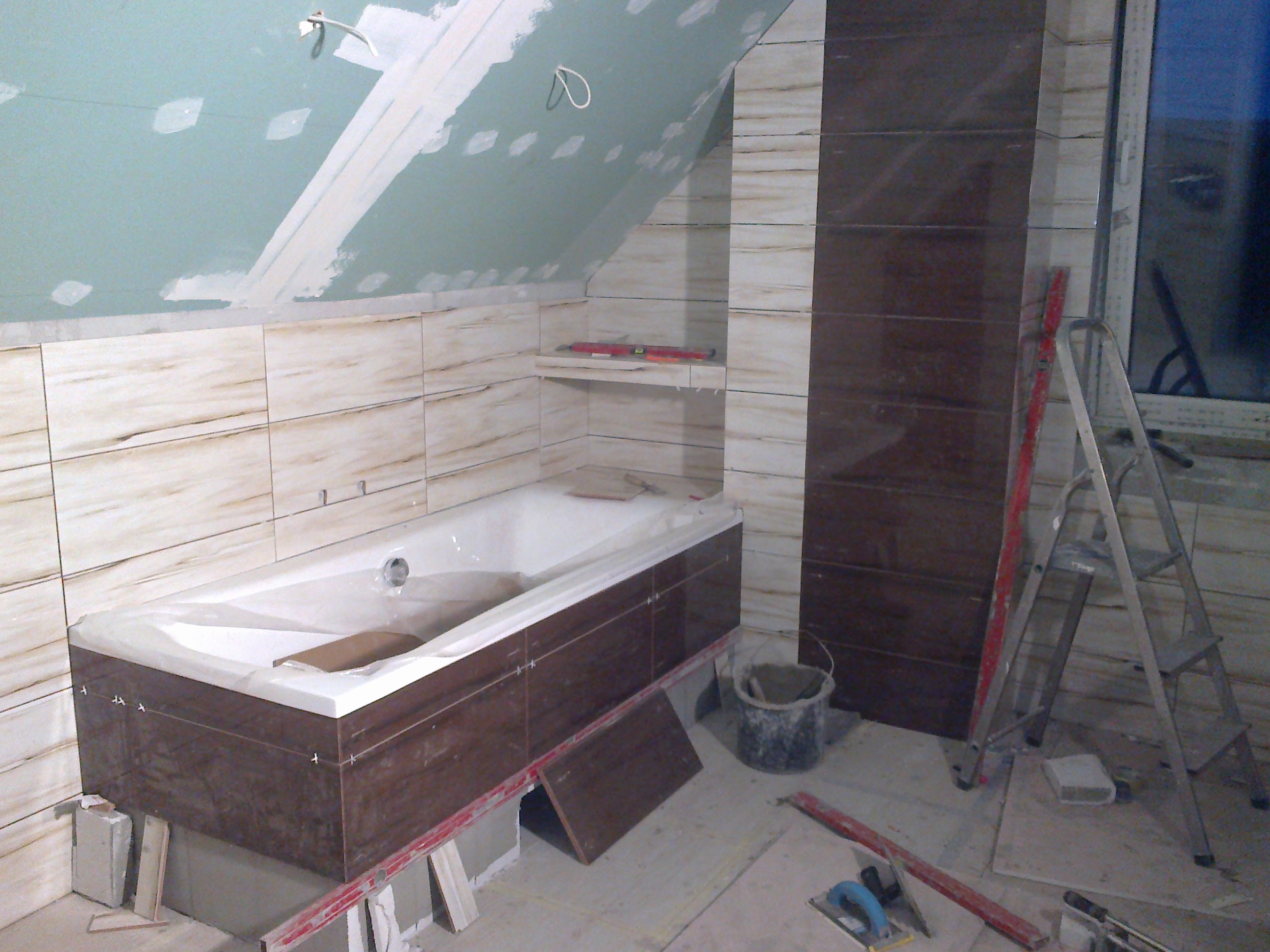 Łazienka w trakcie remontu na poddaszu: obudowana wanna z brązowymi płytkami, ściany wyłożone jasnymi płytkami imitującymi drewno, sufit z płyt kartonowo-gipsowych przed malowaniem, drabina oparta...