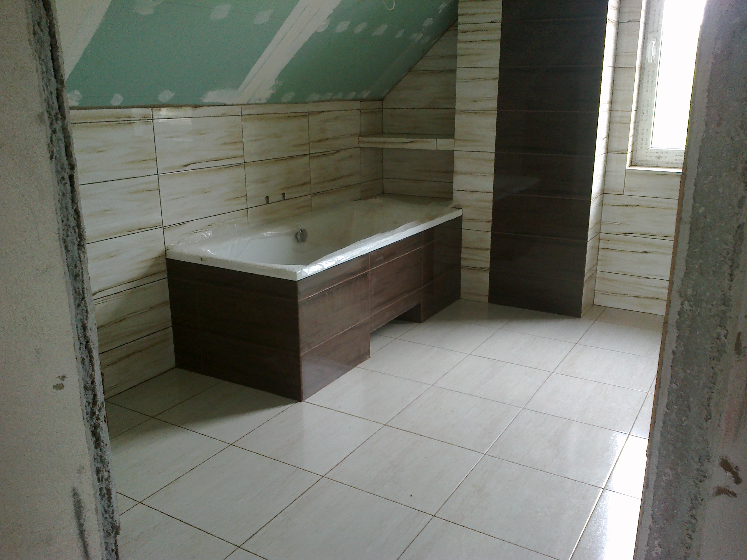 Łazienka w trakcie remontu na poddaszu, z obudowaną brązowymi płytkami wanną, jasnymi płytkami na ścianach i podłodze, oraz surowym wykończeniem ścian i sufitu