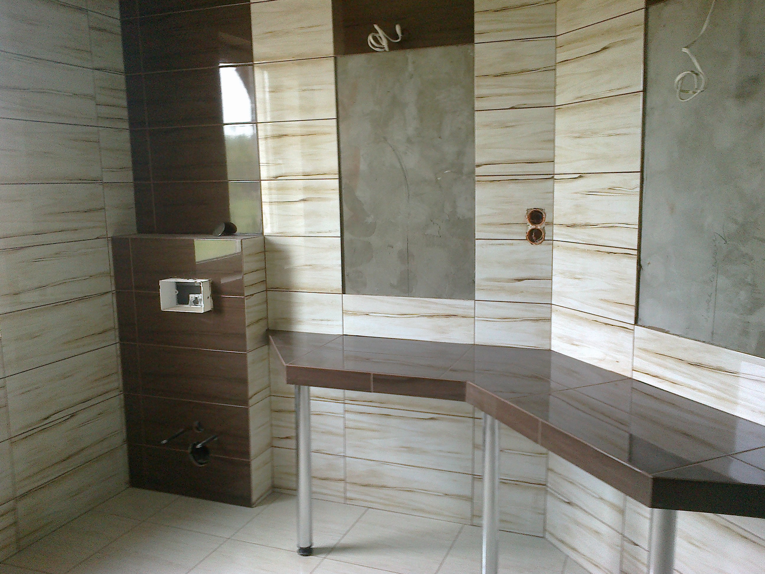 Wnętrze w trakcie wykańczania: kremowe i brązowe płytki imitujące drewno na ścianach, niedokończona ściana z widocznymi przewodami, blat z ciemnych płytek na metalowych nogach.