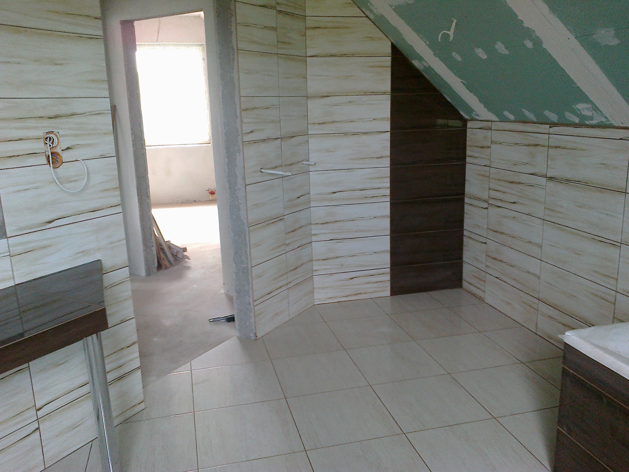 Wnętrze łazienki w trakcie remontu, z beżowymi płytkami imitującymi drewno na ścianach i podłodze, widoczne fragmenty instalacji elektrycznej, surowy sufit pod skosem z płyt gipsowo-kartonowych...