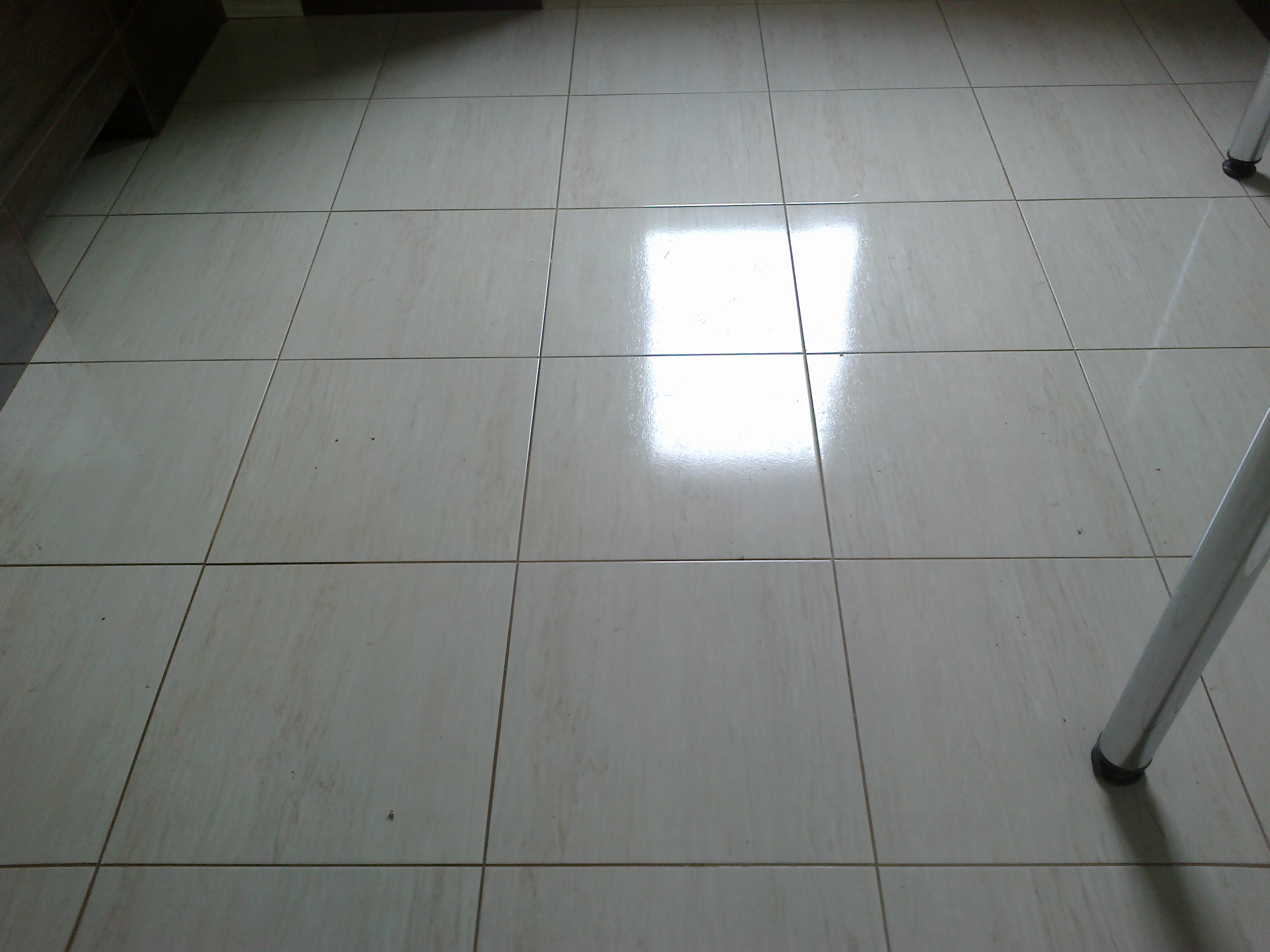 Beżowe, błyszczące płytki podłogowe ułożone w kwadraty, z widocznym odbiciem światła i fragmentem nogi meblowej.