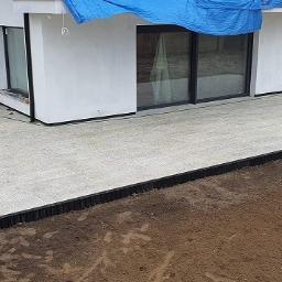 OLBA Bruk - Jasnoszara kostka brukowa ułożona na tarasie domu jednorodzinnego, widoczne czarne obrzeża, pracownicy w odblaskowych kamizelkach w tle.