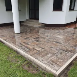 Nowo ułożona, mokra kostka brukowa w odcieniach brązu i szarości, otoczona obrzeżem, przylegająca do białego budynku z kolumną.