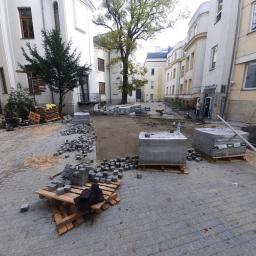 OLBA Bruk - Plac wyłożony szarą kostką brukową w trakcie prac, palety z kostką, przygotowany teren, budynki w tle, widoczny fragment maszyny budowlanej.