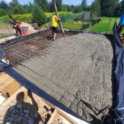 Wylewanie betonu na przygotowane zbrojenie pod fundament, widoczna pompa do betonu, pracownicy w trakcie pracy, teren budowy z zielonym otoczeniem i ogrodzeniem.