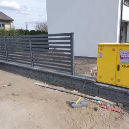 OLBA Bruk - Szare, poziome ogrodzenie panelowe na podmurówce z szarej kostki, żółta skrzynka gazowa, teren w trakcie prac wykończeniowych, widoczny młotek i poziomica.