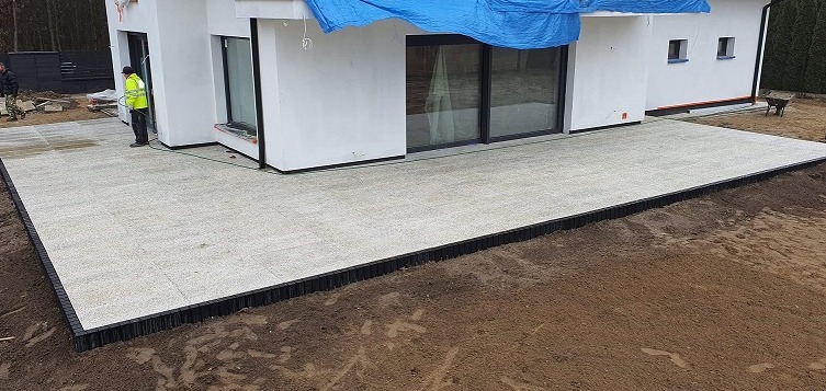 Jasnoszara kostka brukowa ułożona na tarasie domu jednorodzinnego, widoczne czarne obrzeża, pracownicy w odblaskowych kamizelkach w tle.