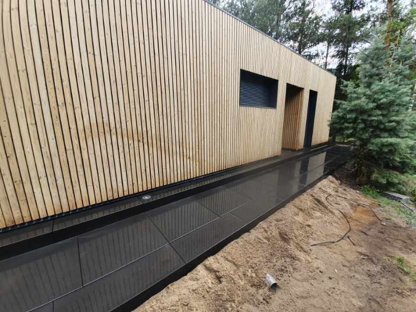 Nowoczesny dom z elewacją z pionowych desek i ciemną, błyszczącą ścieżką z płyt, częściowo otoczoną piaskiem.
