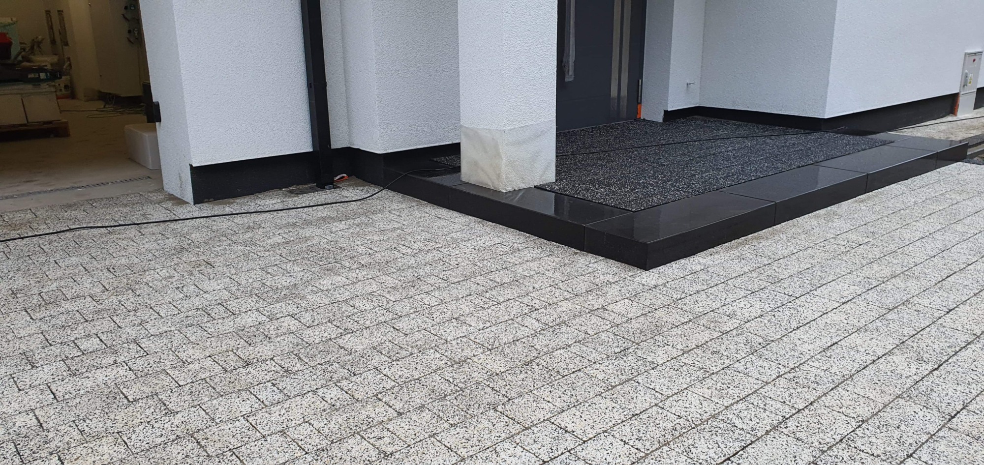 Jasnoszara kostka brukowa ułożona przed wejściem do budynku z czarnymi obramowaniami i wycieraczką.