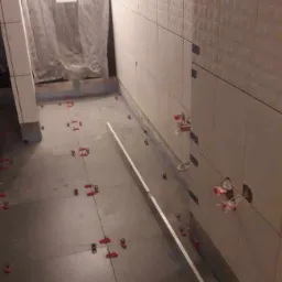 Układanie białych, strukturalnych płytek ściennych w łazience, widoczne krzyżyki dystansowe i poziomica, zabezpieczona folią strefa prysznica.