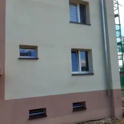 Fragment elewacji budynku po ociepleniu i pomalowaniu na dwa kolory, z widocznymi oknami i rurą spustową, teren wokół budynku częściowo uporządkowany.