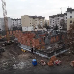 Budowa domu wielorodzinnego w trakcie wznoszenia ścian z cegły, widoczny żuraw budowlany na tle zabudowy miejskiej.