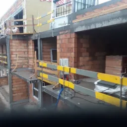 Wznoszenie murów z czerwonej cegły na budowie domu wielorodzinnego z widocznymi elementami szalunków i zabezpieczeń na wysokościach.