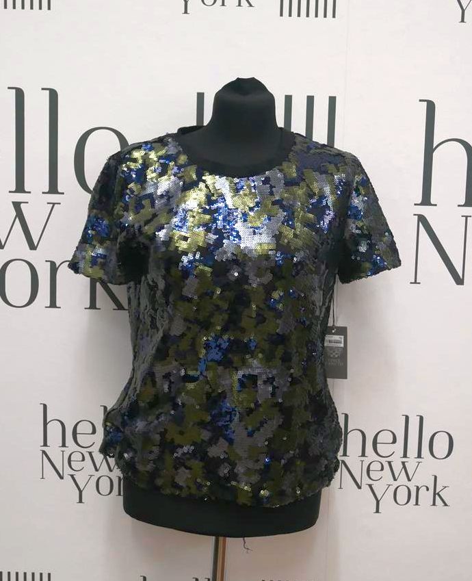 Błyszcząca bluzka z krótkim rękawem, pokryta cekinami w odcieniach zieleni, niebieskiego i szarości, prezentowana na czarnym manekinie na tle ściany z napisem 'hello New York'.