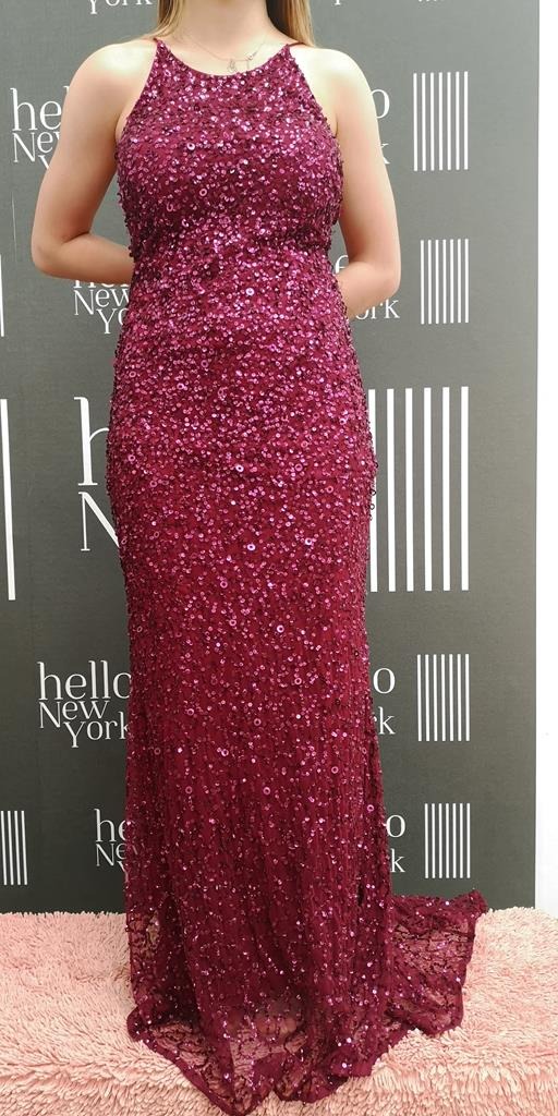Długa, bordowa suknia wieczorowa pokryta cekinami, prezentowana na tle ściany z napisem 'hello New York' i pionowymi paskami.