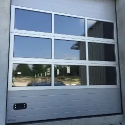 Szara segmentowa brama garażowa z sześcioma prostokątnymi oknami, odbijającymi krajobraz z drzewami i budynkami.