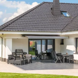 Nowoczesny dom jednorodzinny z tarasem i rozłożoną markizą przeciwsłoneczną, widok z zewnątrz na czarne ramy okien i drzwi tarasowych.