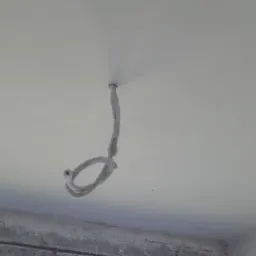 Fragment sufitu z wystającym przewodem elektrycznym, zabezpieczonym białym materiałem, widoczny fragment okna i surowy mur w tle.