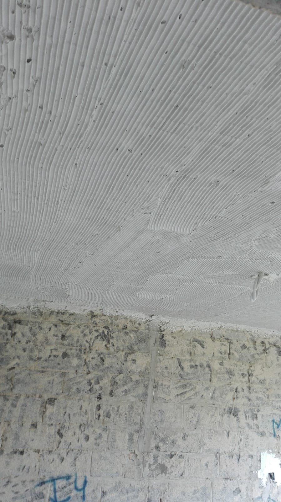 Świeżo nałożony, biały tynk z wyraźnymi, równoległymi śladami po narzędziu, widoczne niedoskonałości i nierówności na powierzchni, surowy mur w dolnej części kadru.