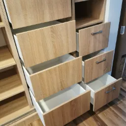 Fragment szafy przesuwnej z otwartymi szufladami o frontach w kolorze jasnego drewna i białych wnętrzach, widoczne metalowe uchwyty i drewniana podłoga.