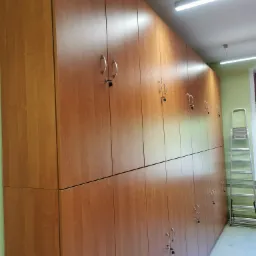 Długa ściana szafek z drewna o jednolitym wzorze, każda z zamkiem i srebrnym uchwytem, ustawiona w rzędzie wzdłuż ściany w jasnym pomieszczeniu, z drabiną opartą o ścianę w tle.