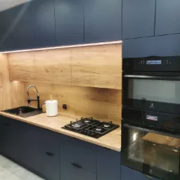 Nowoczesna kuchnia z ciemnoniebieskimi szafkami, drewnianym blatem i panelem nad blatem, z czarnym zlewem, baterią i płytą gazową, oświetlona listwą LED pod szafkami wiszącymi, z piekarnikiem...