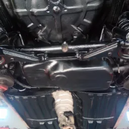 Podwozie samochodu po zabezpieczeniu antykorozyjnym, widoczne elementy zawieszenia, zbiornik paliwa i układ wydechowy zabezpieczone czarną powłoką, auto na podnośniku.