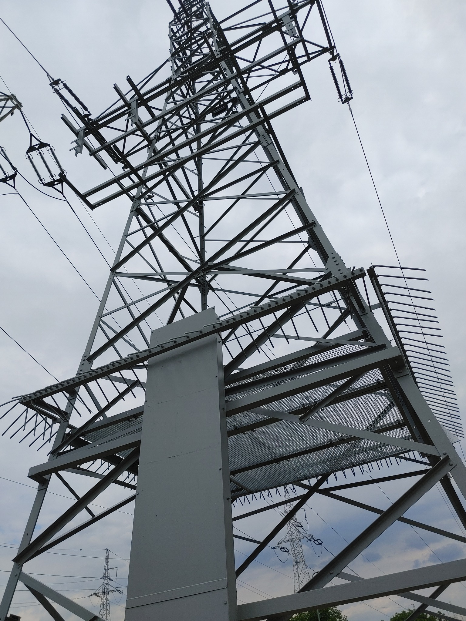 Wysoka, metalowa wieża energetyczna z widoczną konstrukcją kratownicową i przewodami, sfotografowana z dołu na tle pochmurnego nieba. Widoczne elementy anty-ptasie na górze konstrukcji.