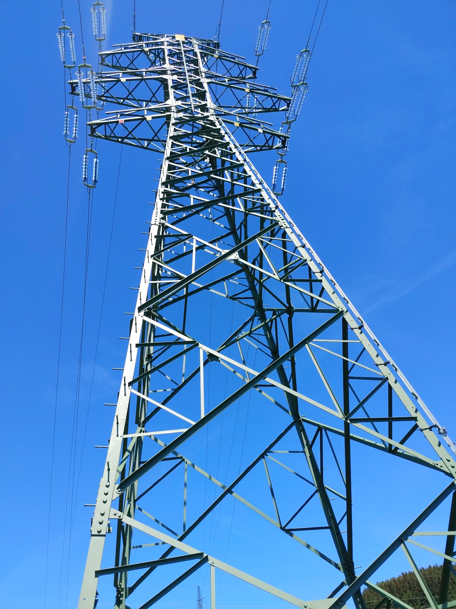 Wysoki słup wysokiego napięcia o konstrukcji kratownicowej, pomalowany na zielono, widoczny z dołu na tle błękitnego nieba, z widocznymi izolatorami i przewodami.