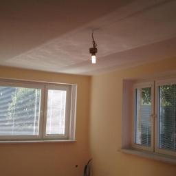 Fixer Company - Pokój po szpachlowaniu z dwoma oknami w białych ramach i żarówką zwisającą z sufitu. Ściany w odcieniach beżu, przygotowane pod malowanie.