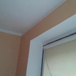 Fixer Company - Pomarańczowa ściana z białym sufitem i listwą dekoracyjną oraz biała rama okienna z roletą. Widok na róg pokoju.